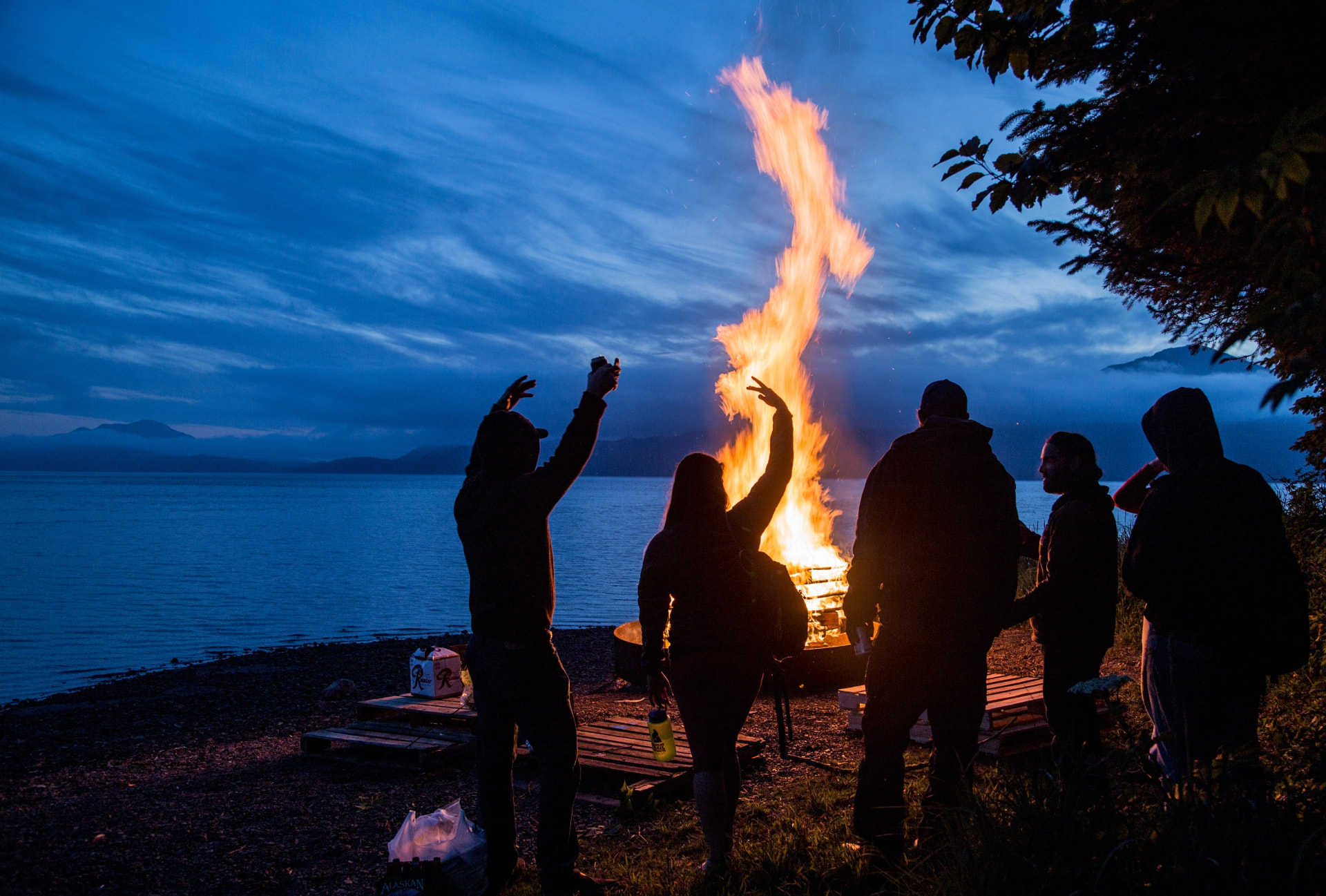 Friends gathered around a tall waterfront bonfire at dusk.